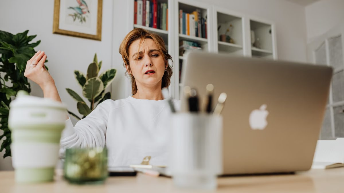 A woman in a home office setting works on a laptop, surrounded by books and plants.